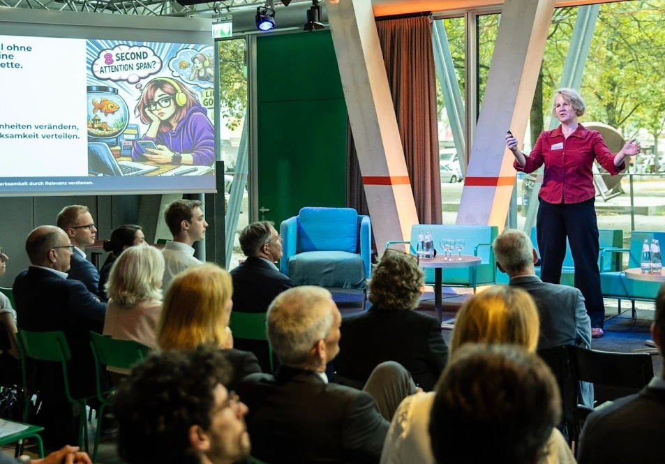 Yvonne Behnke in roter Bluse auf einer hellen Bühne, mit Leinwand und Publikum.